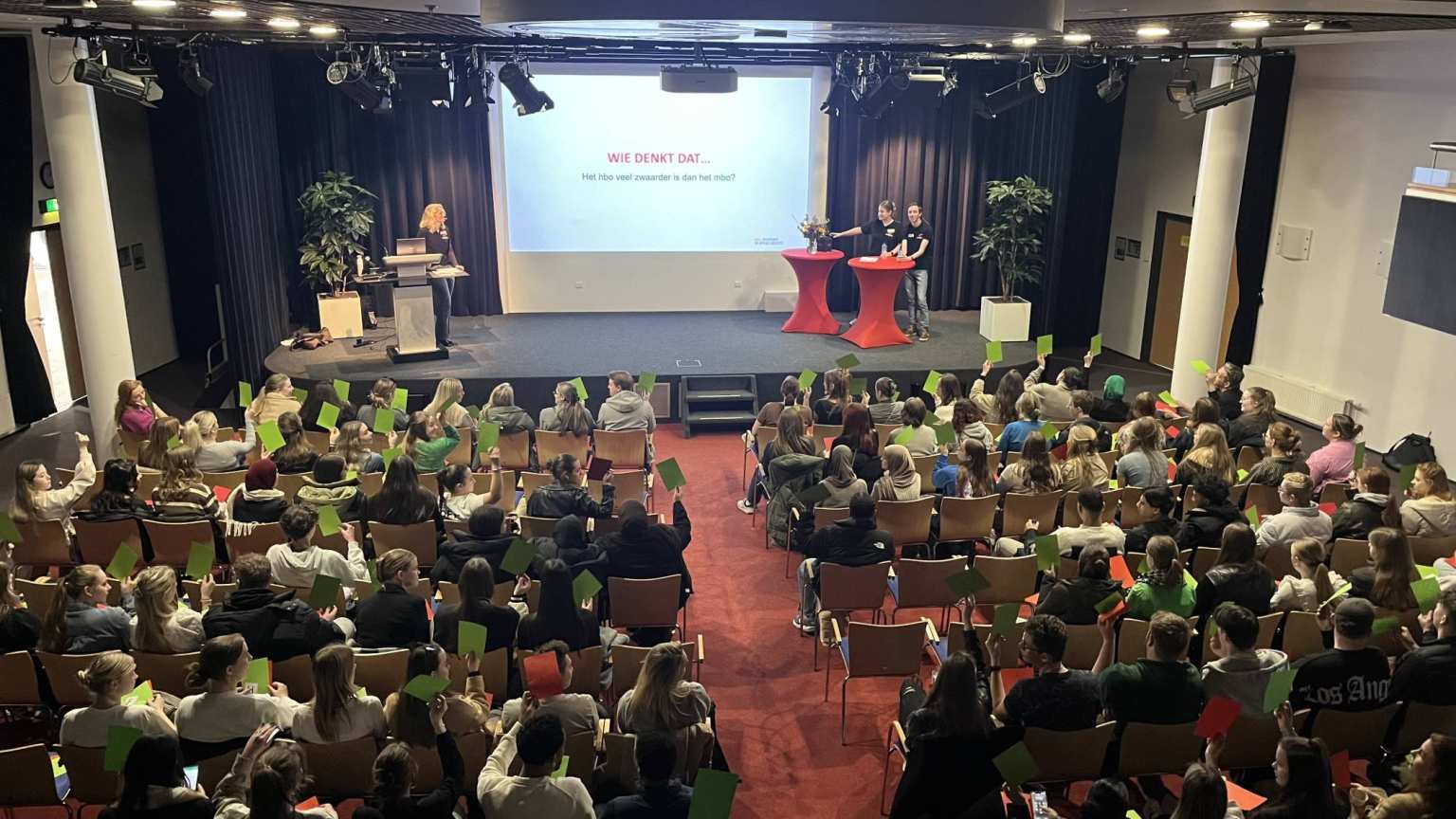 Foto's van de plenaire sessie met mbo-scholieren die groene kaartjes omhoog houden, voor de mbo-hbo proefstudeermiddag van Educatie en Mens en Maatschappij op 7 maart 2024