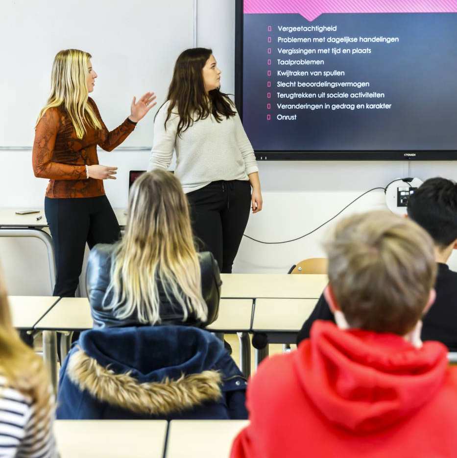 2 vrouwelijke studenten Social Work staan voor de klas en geven een presentatie. Je leert heel veel kennis en vaardigheden.