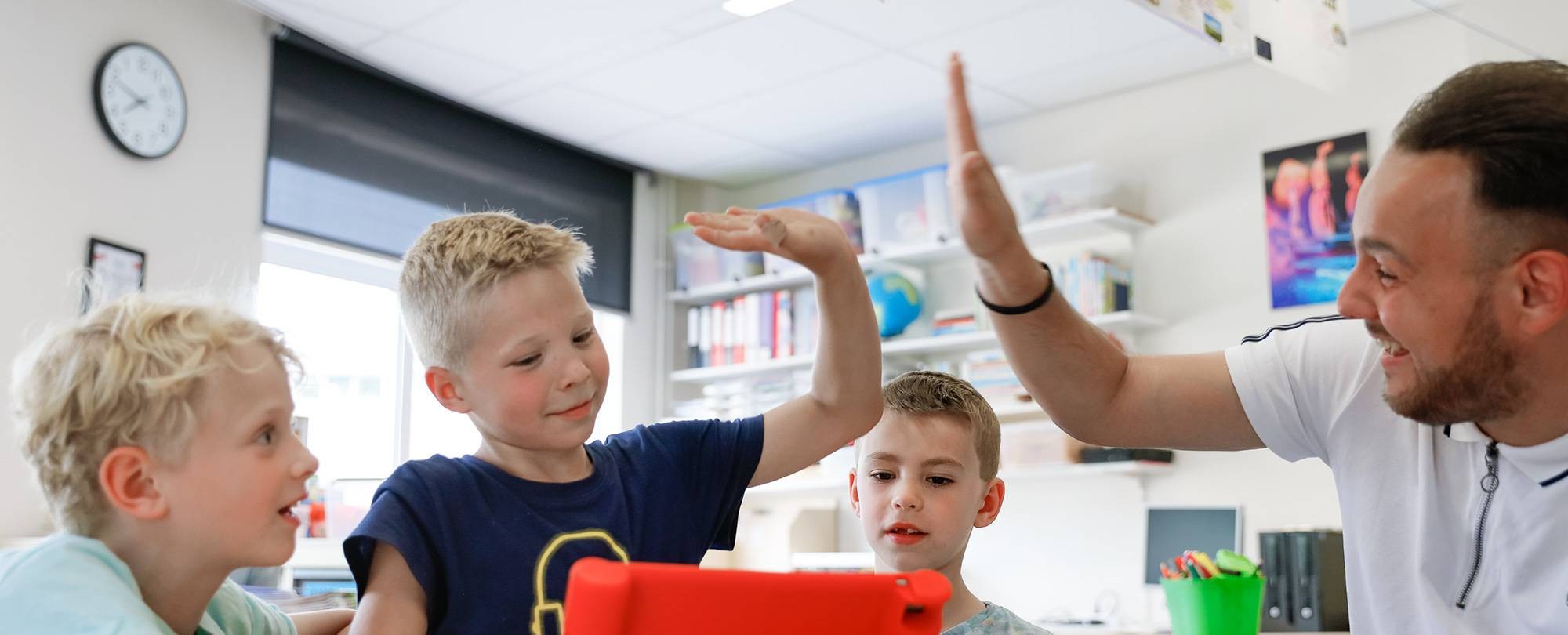 Leraar zit met drie kinderen achter de iPad en geeft n van die leerlingen een high-five.