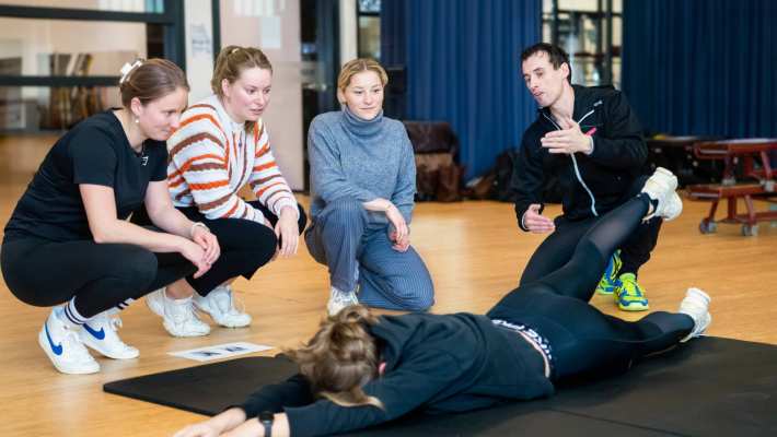 Student ligt op haar buik op de grond, heft een van haar benen de lucht in. Docent en 3 studenten kijken toe.