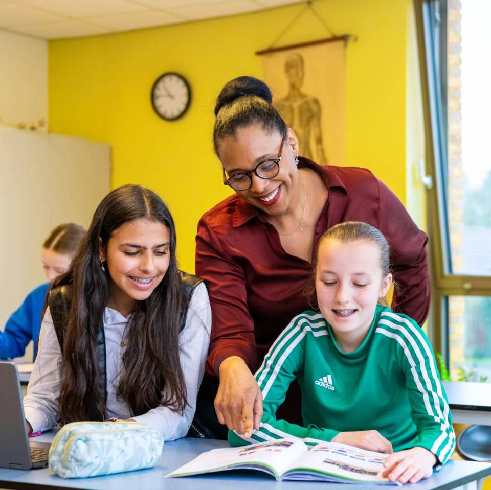 2 lachende leerlingen kijken naar een boek waar de docenten heen wijst op het Citadel College.