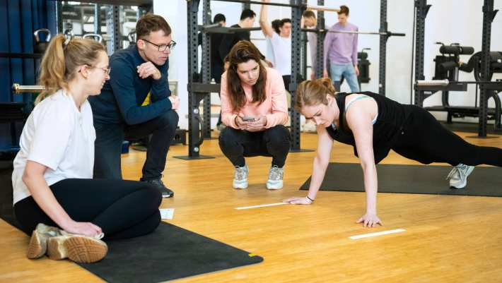 Student staat in een plank positie, 3 studenten kijken toe.