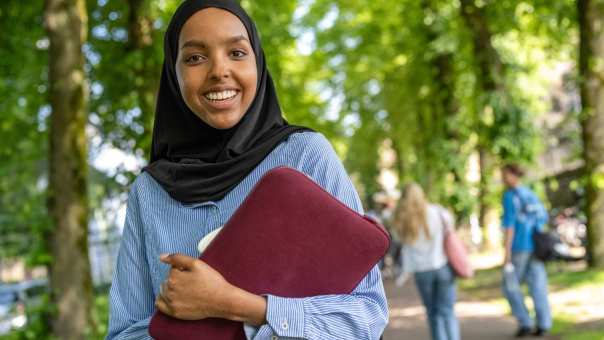 Studente met hoofddoek, loopt buiten en kijkt in de camera, heeft een laptop vast. 