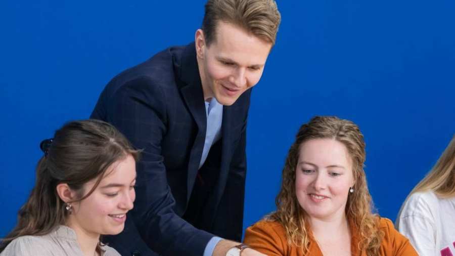 Docent in donkerblauw pak kijkt mee op de laptop bij twee van zijn studenten.
