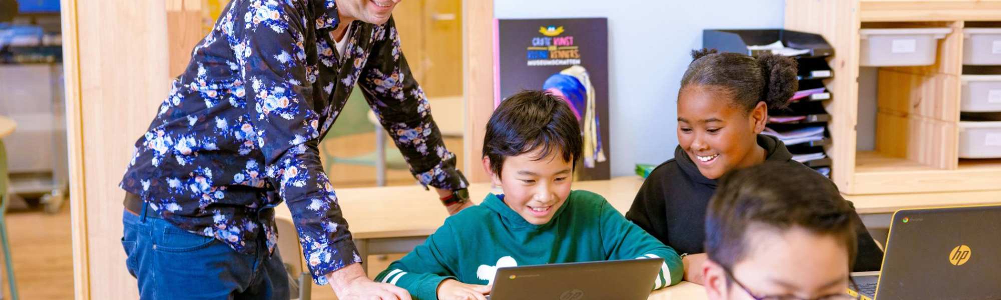 Groep 5,6,7 op de school Da Vince. De leraar en twee leerlingen lachen die achter hun laptop aan het werk zijn. 