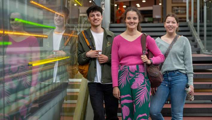 Drie studenten lachen in de camera, staan op de trap in lichtstraat van gebouw R26. 