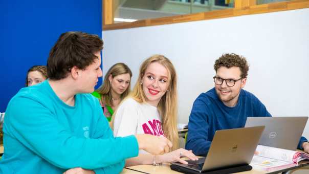 Drie studenten zitten op een rijtje in het klaslokaal met hun laptops opengeklapt. Ze zijn aan het overleggen.
