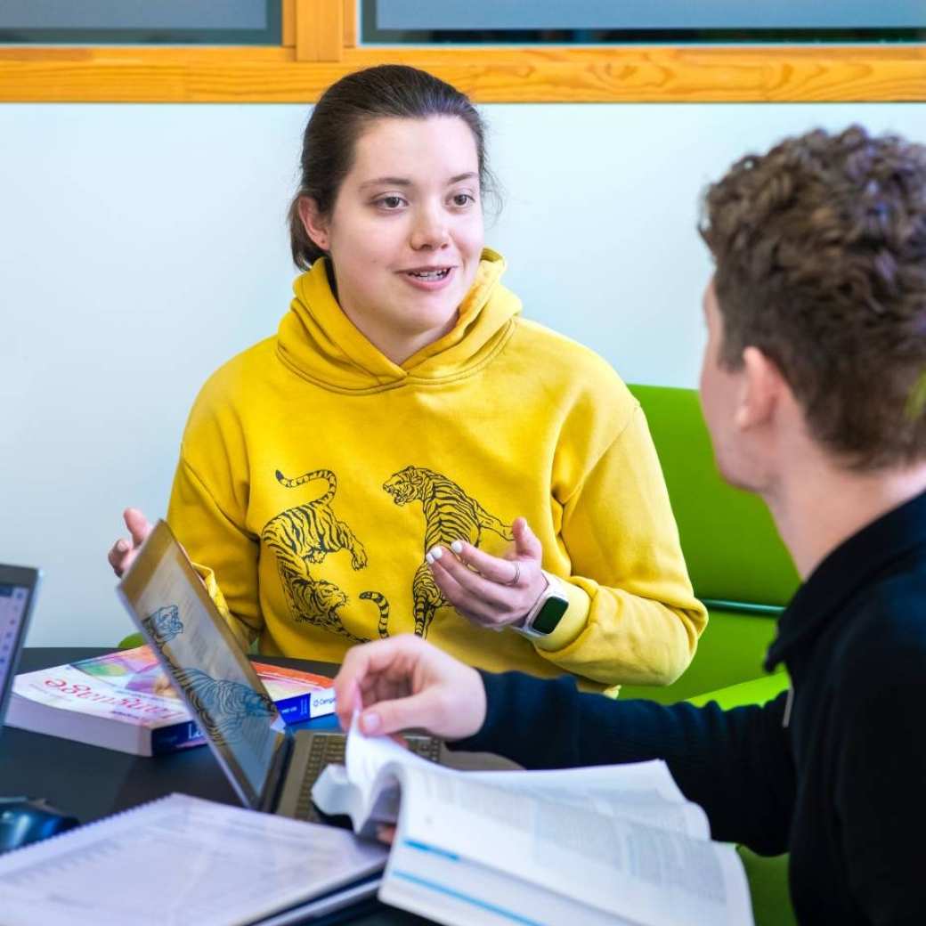 Foto Master Academie Educatie. 2 masterstudenten zijn met elkaar in gesprek aan een tafel met laptops en boeken. 