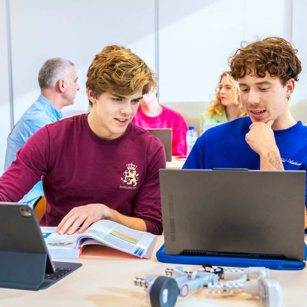 2 mannelijke studenten overleggen met een boek en 2 computers. 