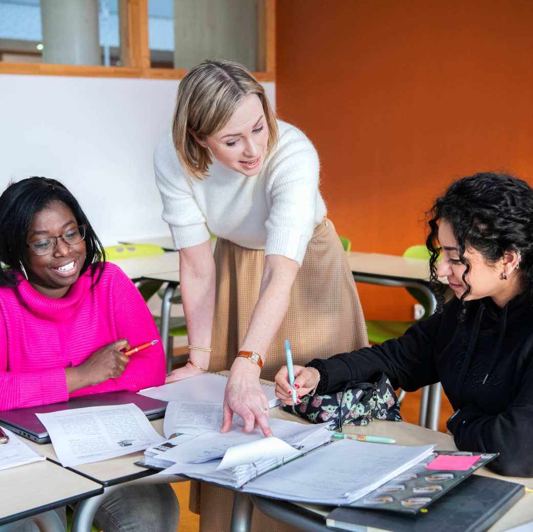 Docent wijst in een boek terwijl 2 vrouwelijke studentes kijken