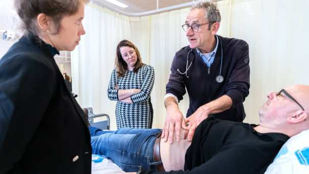 Docent van de HAN Master Physician Assistant geeft uitleg aan 2 studenten over palpatie bij een patint op de onderzoeksbank