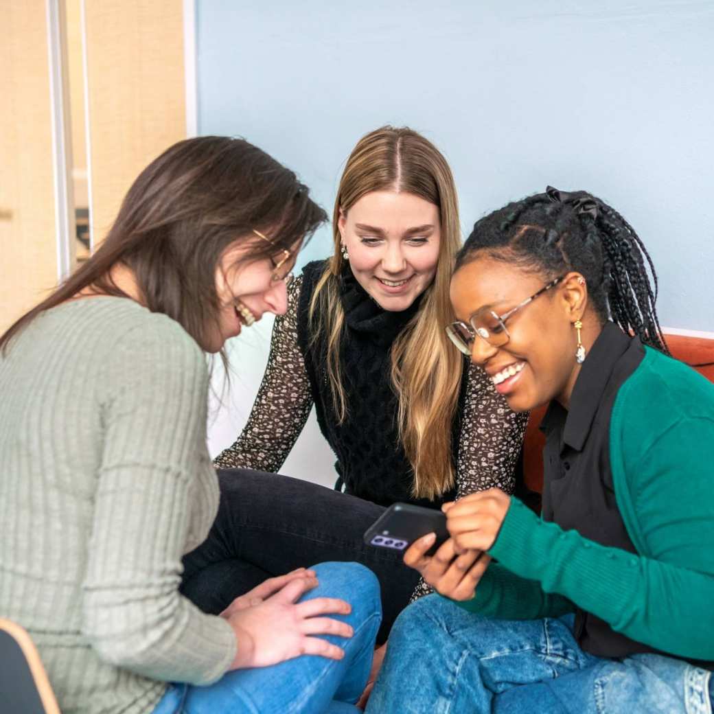 3 studenten zitten samen op een bank en kijken lachend naar een telefoon.