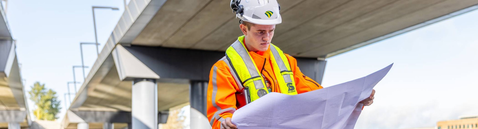 Man met veiligheidshelm en oranje werkjas checkt  de bouwtekening onder een viaduct in aanbouw.