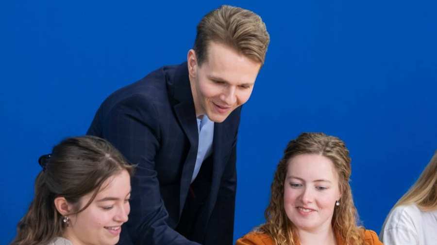 Docent in donkerblauw pak kijkt mee op de laptop bij twee van zijn studenten.
