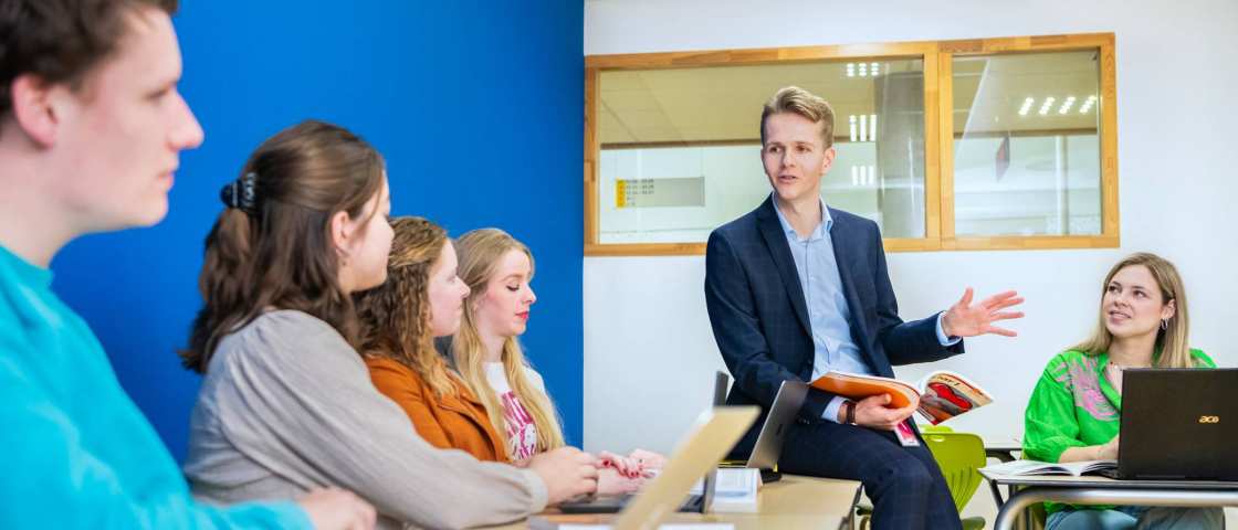 Docent zit op tafel en legt iets uit aan de klas