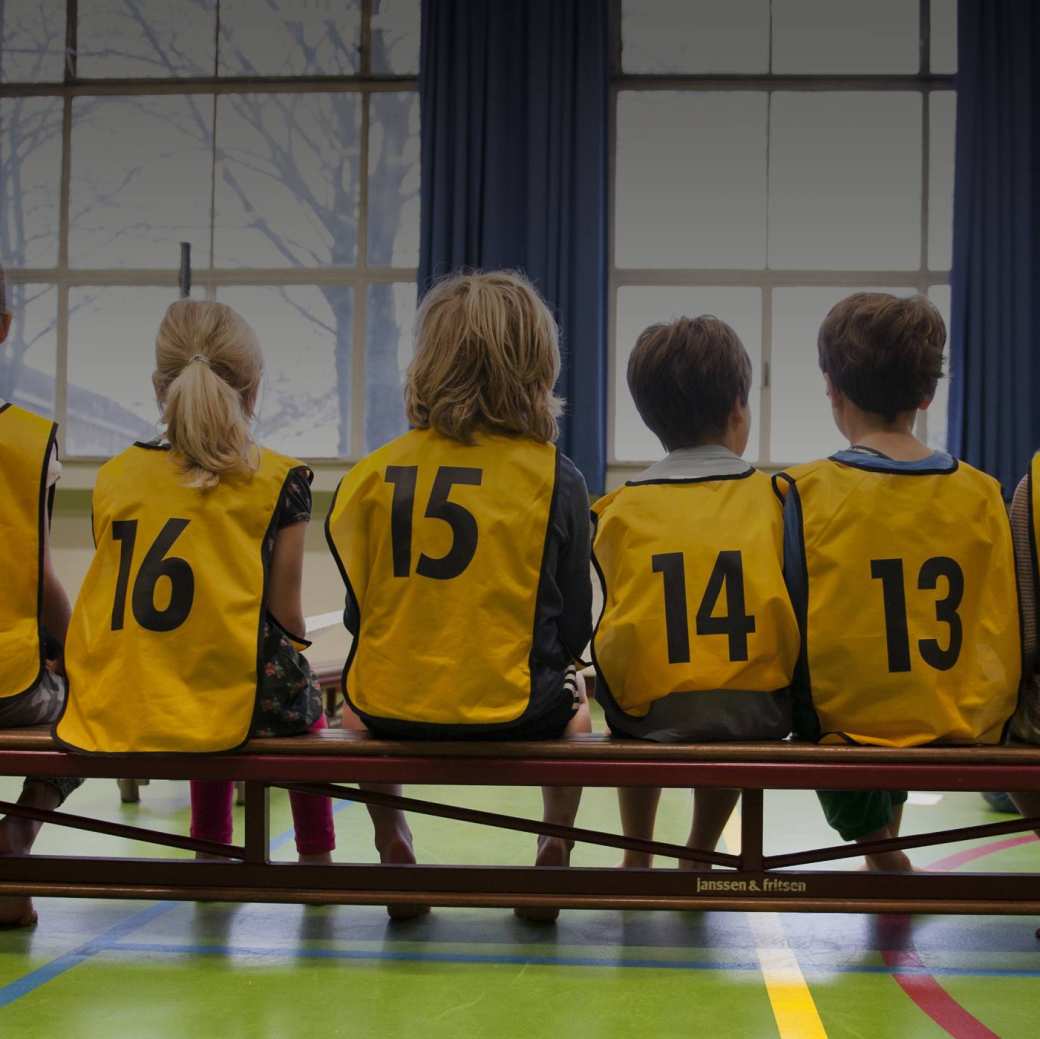 Kinderen in een gymzaal met hesjes aan op de rug gefotografeerd