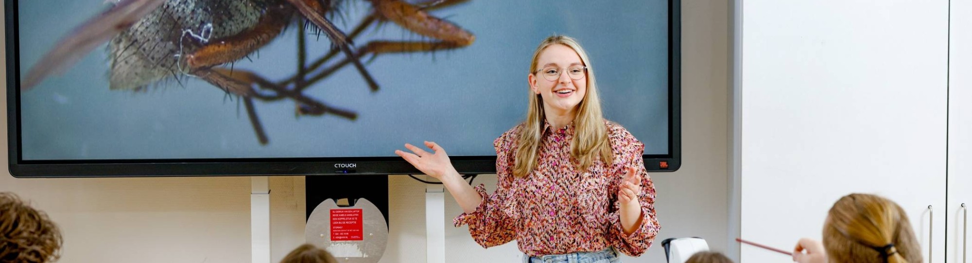 Studente geeft lachend les/staat lachend voor de klas. Op de achtergrond staat een vlieg geprojecteerd.