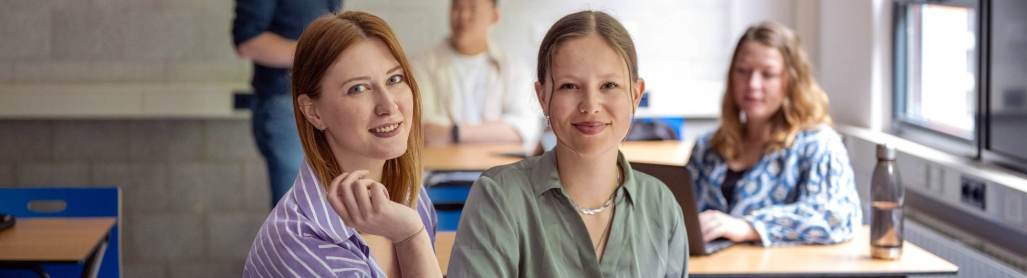 Twee studenten zijn samen aan het werk op de laptop, kijken in de camera, zijn in een klas. 