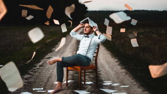 man met wit overhemd, vlinderdas en bretels zit op een stoel, midden op een zandweg. Hi houdt een boek boven zijn hoofd en losse pagina's vliegen in het rond