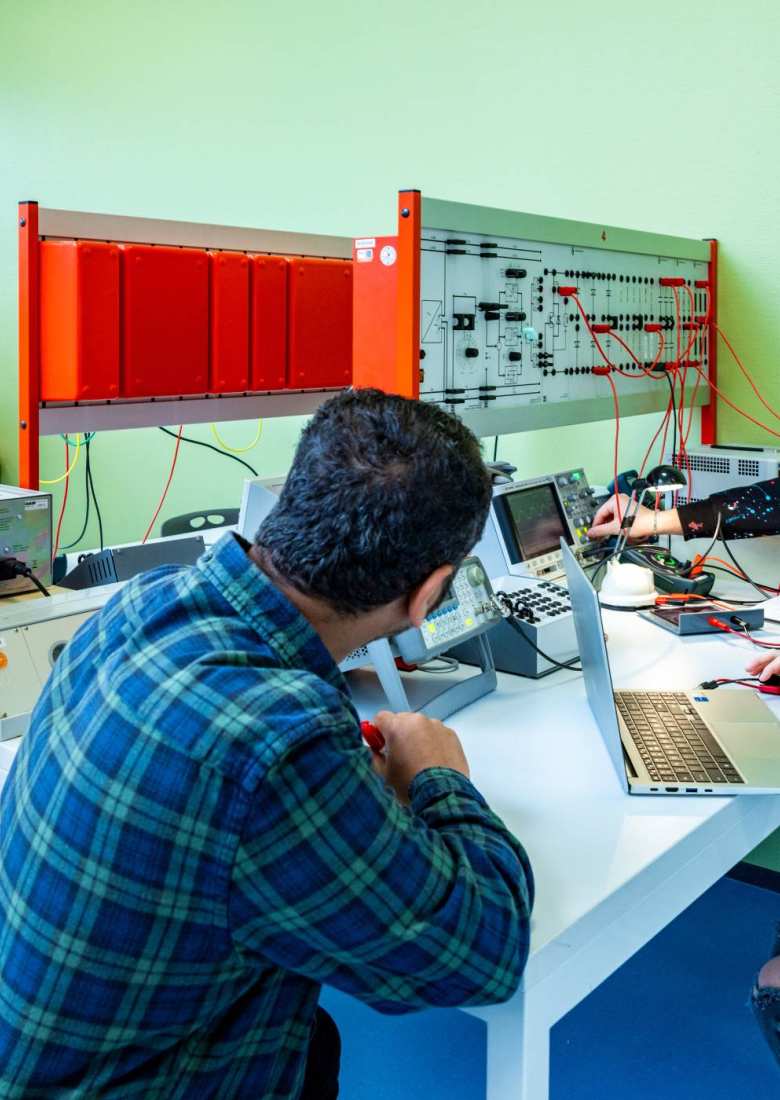 Een elektrotechniekdocent kijkt mee met studenten tijdens practicum.