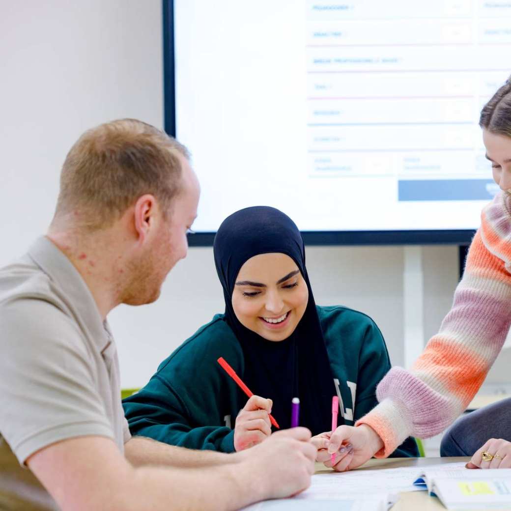 Drie PABO-ALO studenten zitten te werken en één student stelt een vraag aan de docent die aandachtig luistert.