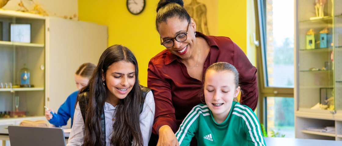 2 lachende leerlingen kijken naar een boek waar de docenten heen wijst op het Citadel College. 