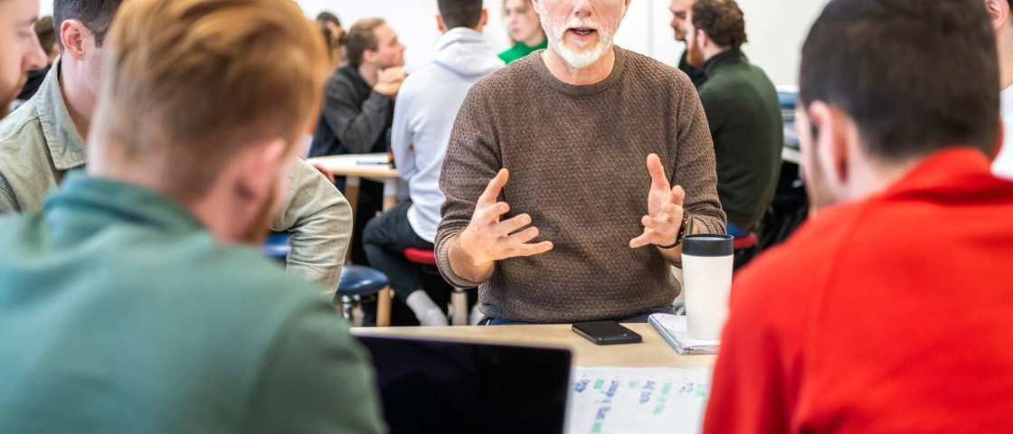 Docent zit bij studenten aan tafel en is aan het woord.