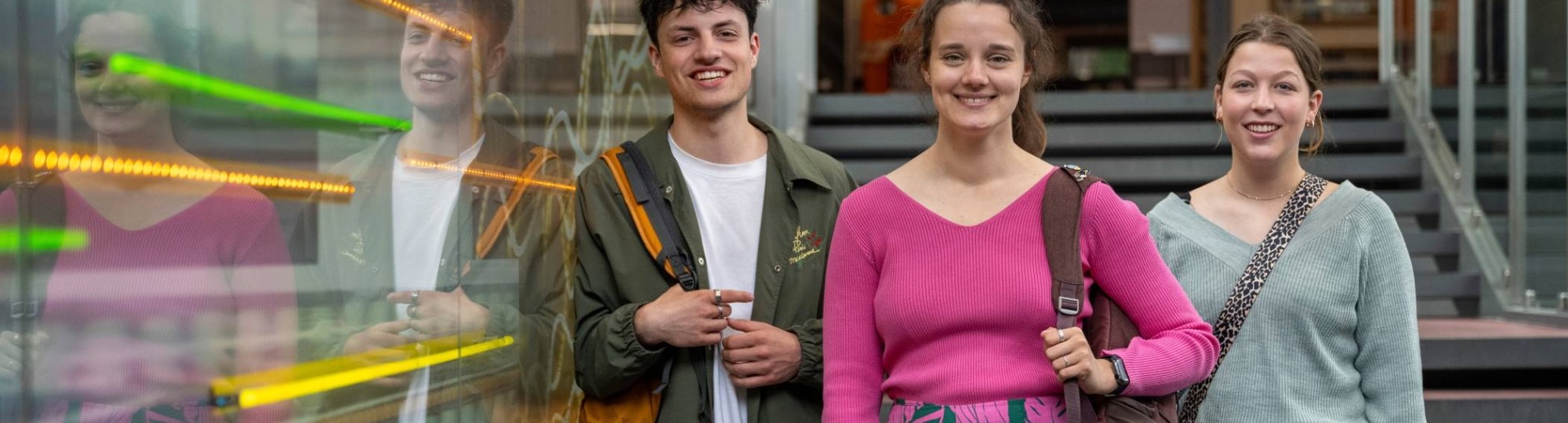 Drie studenten lachen in de camera, staan op de trap in lichtstraat van gebouw R26. 