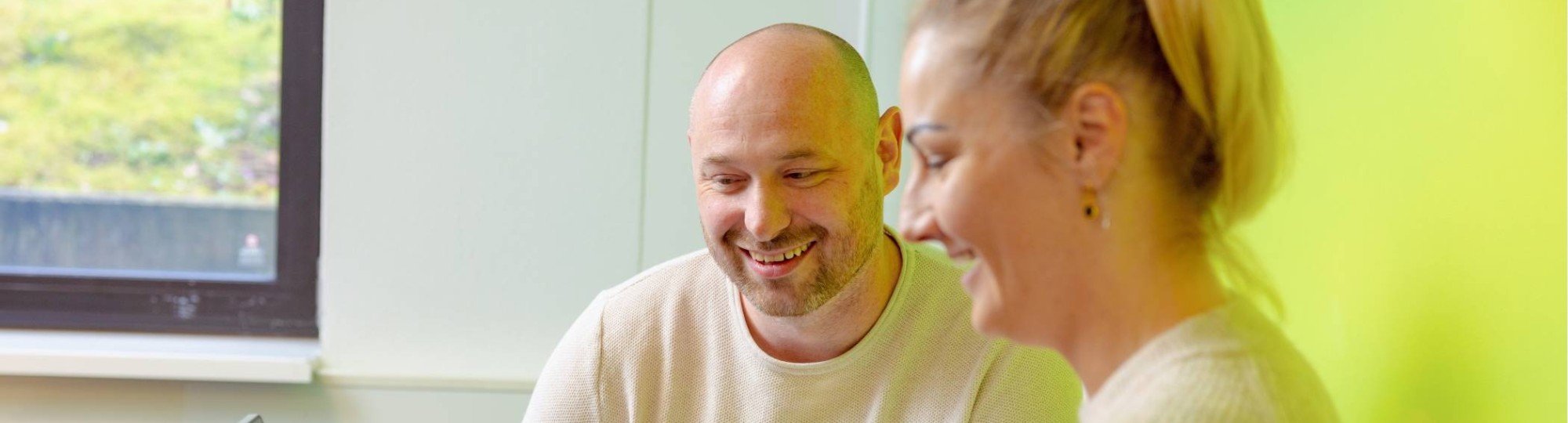 Vrouw en man zitten achter laptop, allebei aan het lachen. Groene muur op achtergrond.