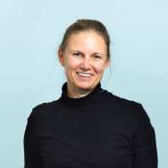 Portrait headshot. The picture is taken in front of a blue background and the subject is wearing a a black turtleneck shirt. Her hair is brunette and tied behind her head. Her eyes are blue. Her right hand is in the right pocket of her beige and brown plaid pleated pants.. She is facing the camera, with shoulders slightly towards the left. 