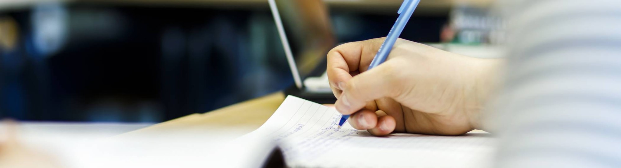 een hand met pen schrijft op een kladblok. erachter staat een laptop en op de voorgrond liggen wat papieren