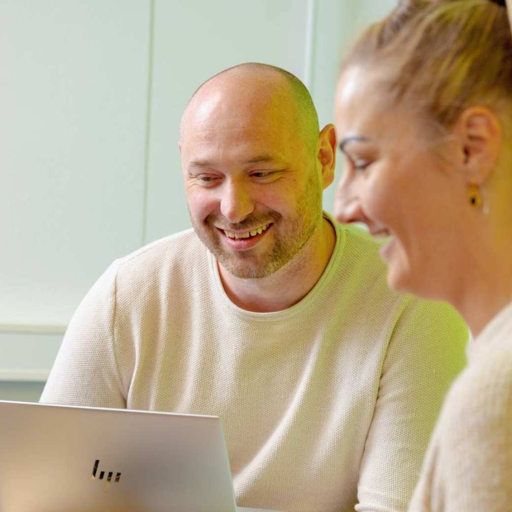 Vrouw en man zitten achter laptop, allebei aan het lachen. Groene muur op achtergrond.