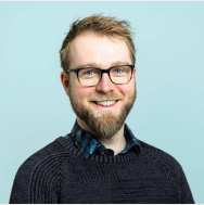 Portrait headshot. The picture is taken in front of a blue background and the subject is wearing a grey knit sweater with a blue and navy collared shirt underneath. His hair is blonde, his eyes are blue-grey, and he has a beard. He wears dark glasses. He is facing the camera, with his shoulders pointing slightly towards the right. 