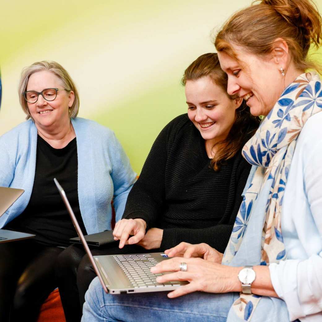 Vrouw wijst iets aan op laptop, oranje banken. Andere vrouw pakt laptop op. Wereldbol op achtergrond.