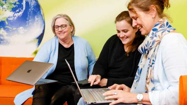 Vrouw wijst iets aan op laptop, oranje banken. Andere vrouw pakt laptop op. Wereldbol op achtergrond.