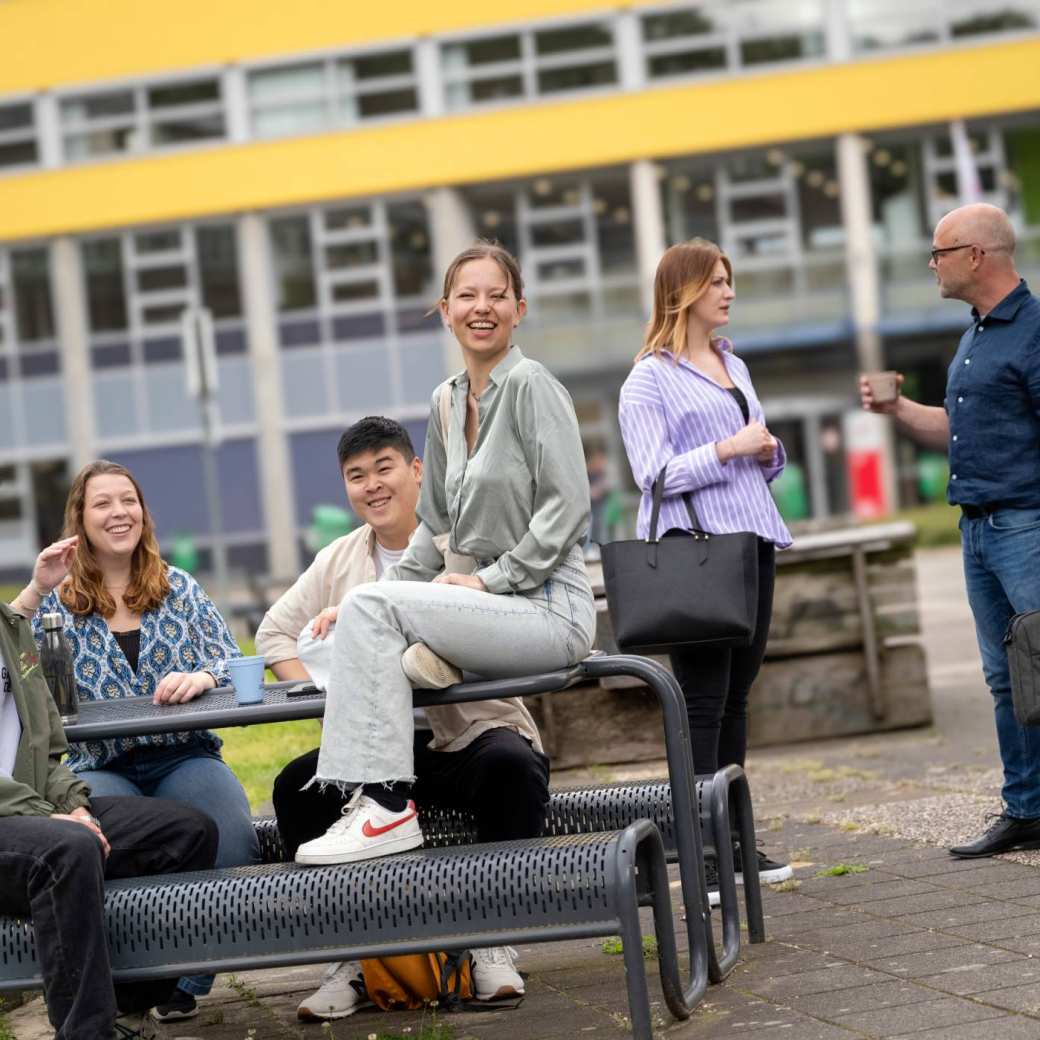 Groepje studenten zitten buiten aan de picknicktafel, op de campus Arnhem, kijken in de camera. 