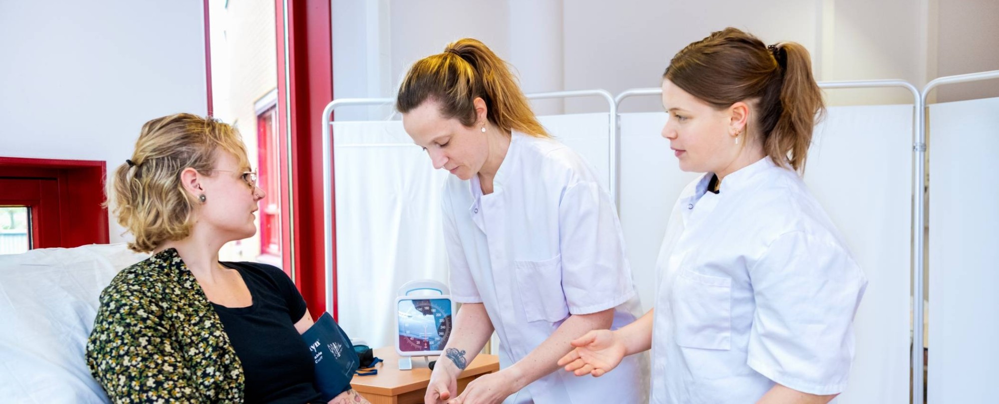 3 studenten oefenen met bloeddruk meten op bed tijdens les Verpleegkunde deeltijd