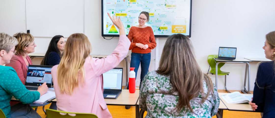 Docent staat voor een volle klas en kijkt lachend naar de student die de vinger opsteekt. 