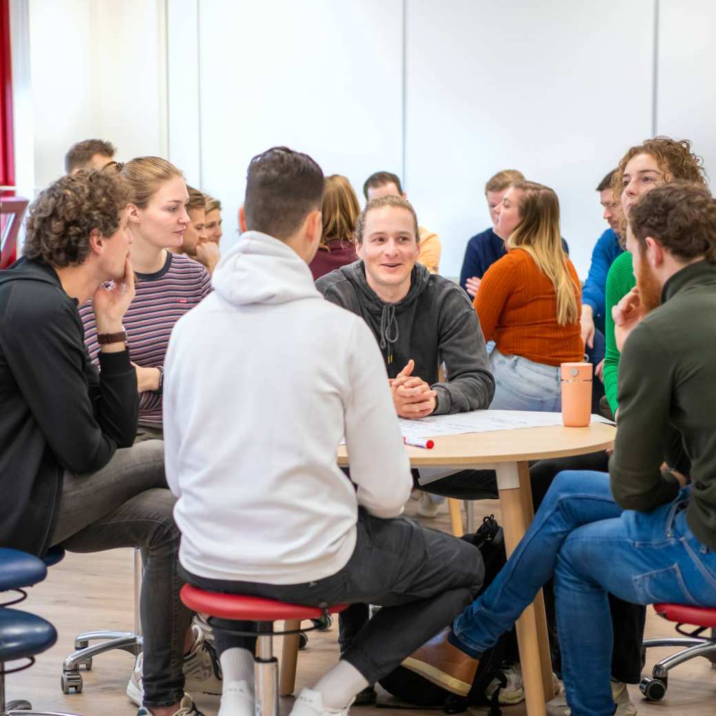 Studenten zitten verdeeld over 2 groepen in een lokaal, kletsend met elkaar.