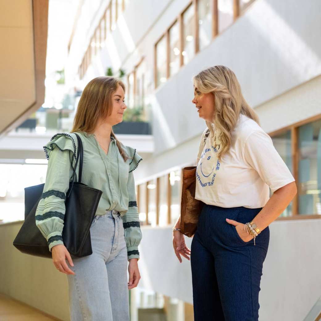 Twee vrouwelijke studenten kletsen met elkaar in de gang van K35 bij de trap. 