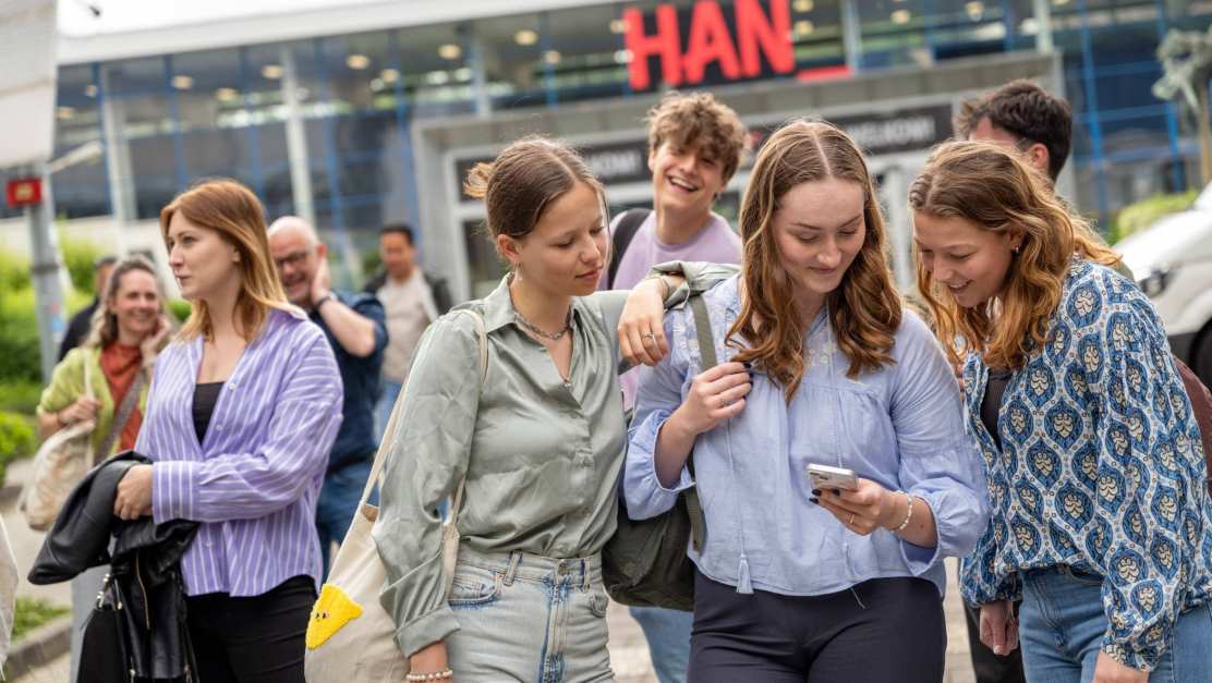 Studenten staan met een groepje buiten bij de ingang van gebouw R26, kijken op een telefoon. 