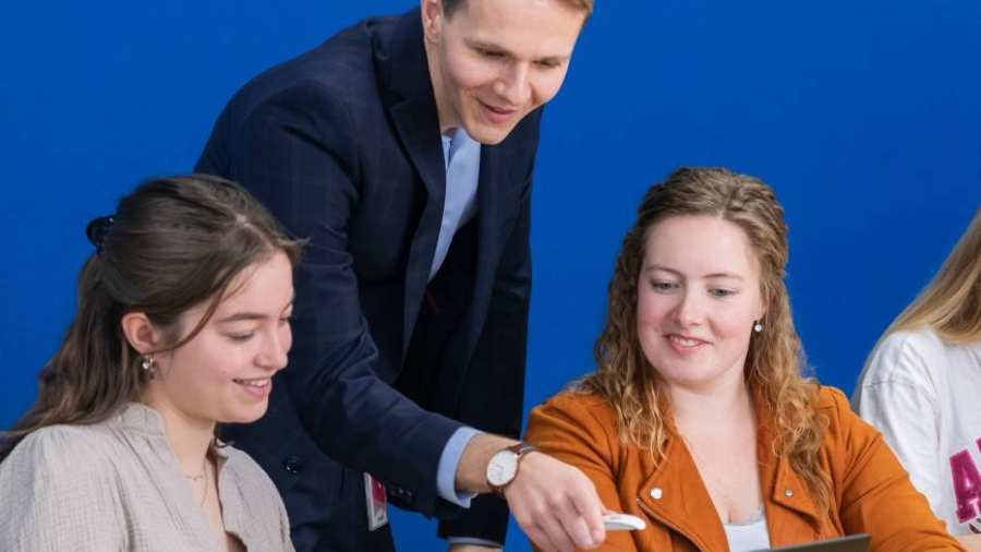 Docent in donkerblauw pak kijkt mee op de laptop bij twee van zijn studenten.
