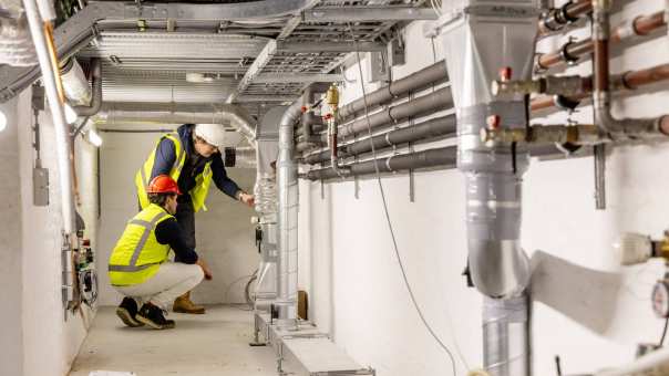 een installatieruimte met leidingen. achterin zit een man gehurkt met een geel hesje en een rode helm.