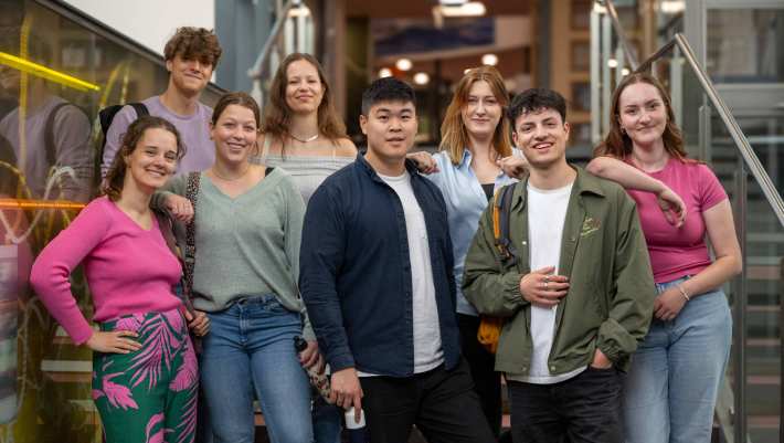 Groep studenten lachen in de camera, staan in de lichtstraat van gebouw R26. 