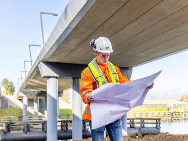 Man met veiligheidshelm en oranje werkjas checkt  de bouwtekening onder een viaduct in aanbouw.