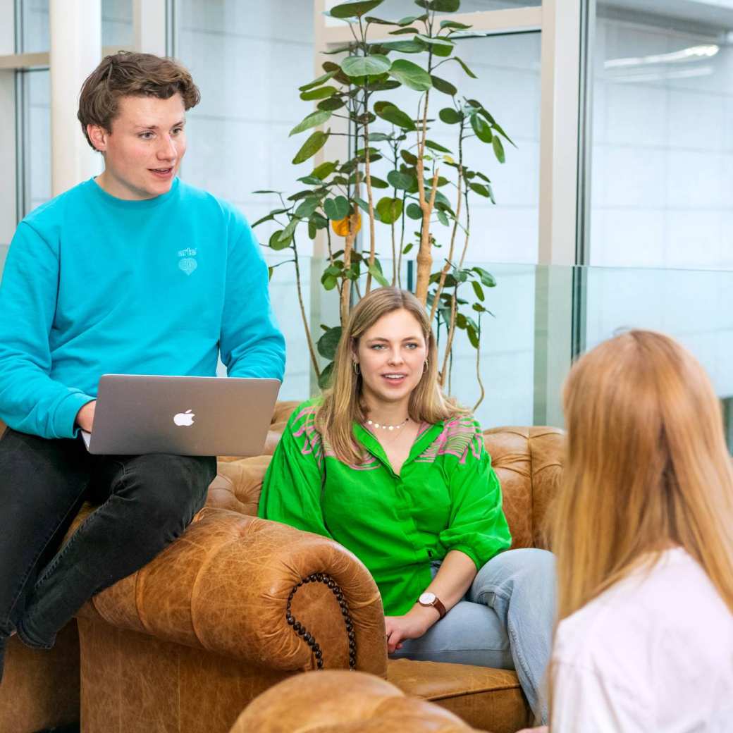 Twee studenten Leraar Duits zitten met hun laptop op schoot op de sofa iets uit te leggen aan een andere student die tegenover hen zit.
