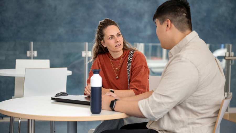 Docent in gesprek met student aan een tafel op podium in hal van gebouw R26, studentbegeleiding.