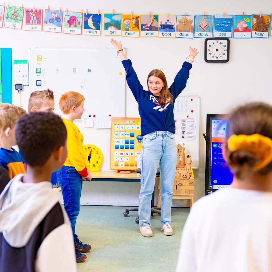 Juf staat voor de klas en heeft handen in de lucht