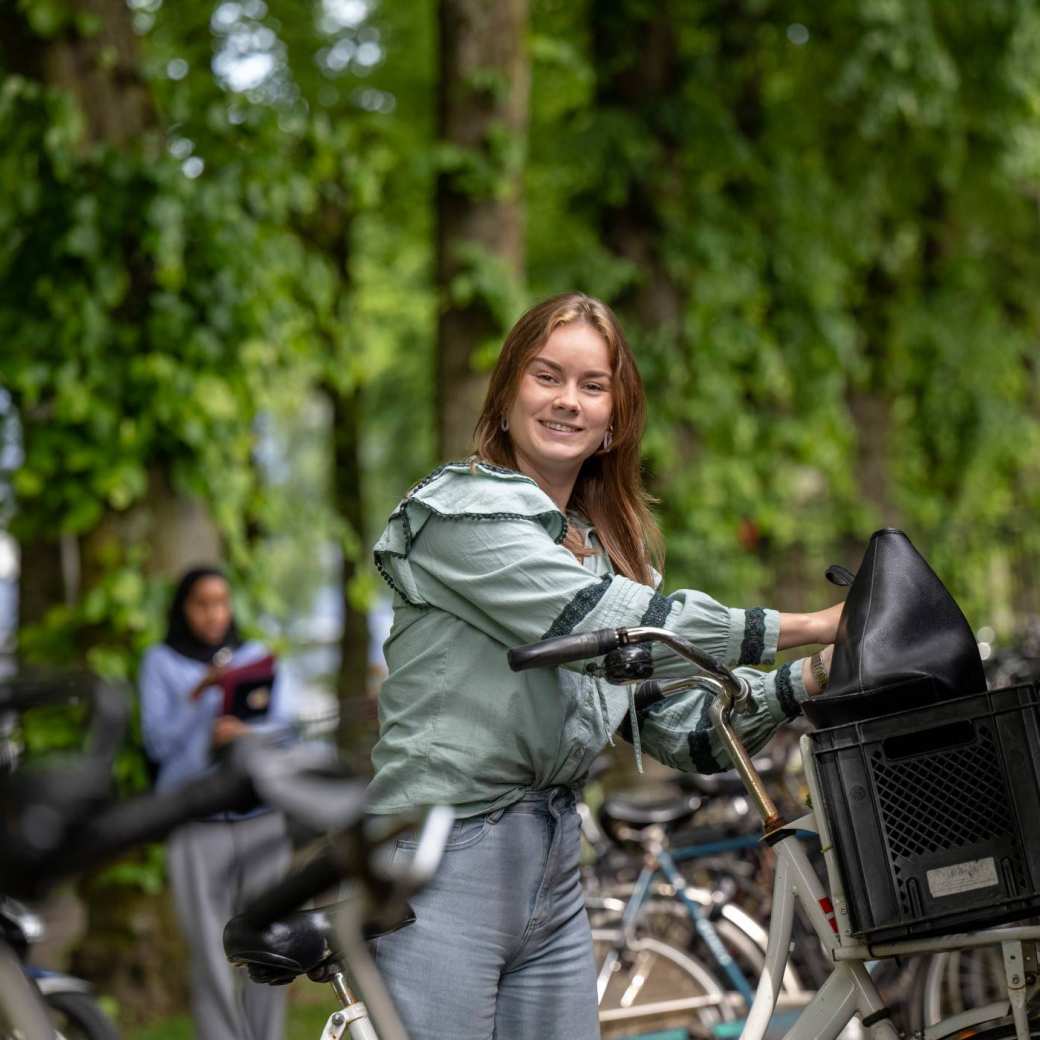 Student parkeert haar fiets in de fietsenstalling bij LvS10, kijkt in de camera, op campusterrein. 