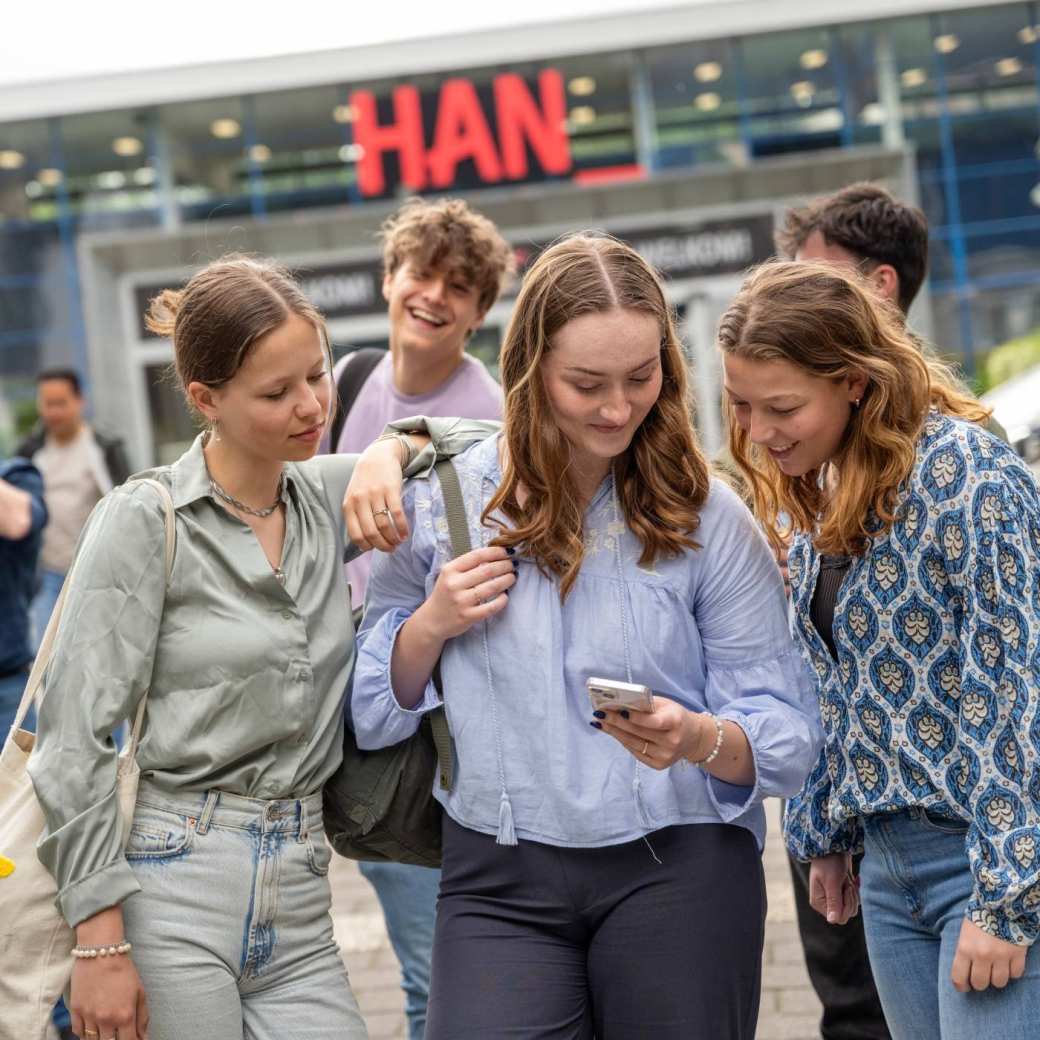 Studenten staan met een groepje buiten bij de ingang van gebouw R26, kijken op een telefoon. 